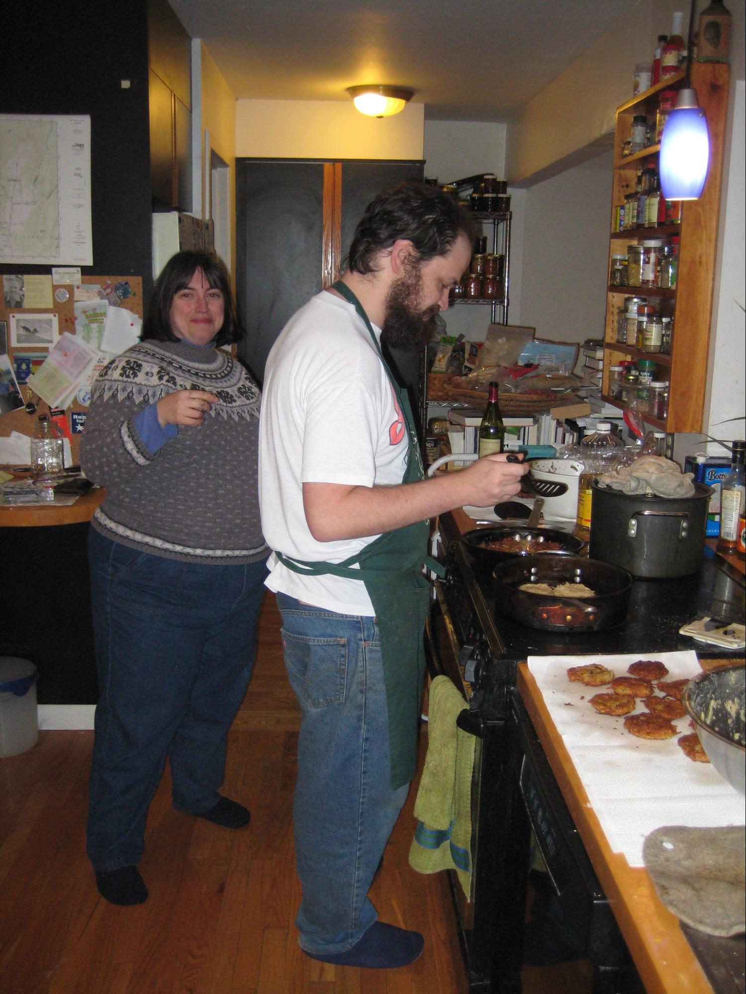 A man in an apron prepares latkes while a woman smiles and looks on