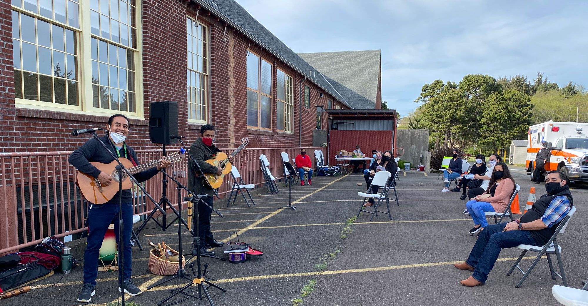 A brick community center provides a backdrop to musicians playing guitars at a COVID-19 vaccination clinic. Vaccine recipients wearing mass sit carefully spaced out to listen. An ambulance and health volunteers are in the background.