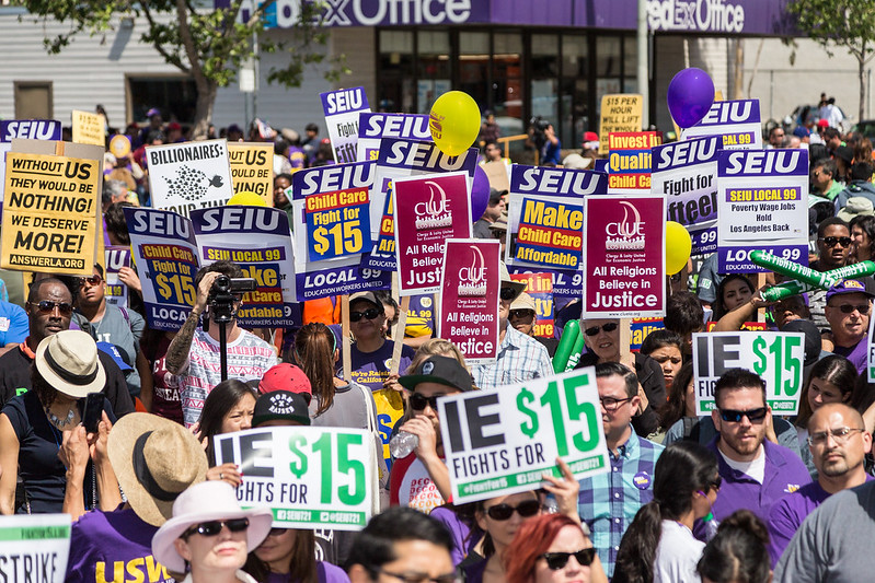 A diverse crowd of people hold signs advocating for a $15 minimum wage on the street in front of a FedEx Office