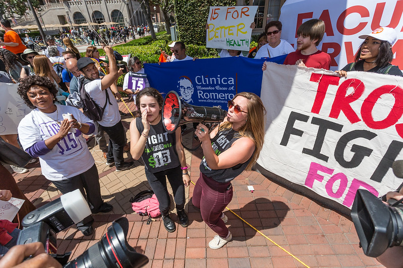 A diverse group of people hold signs, smile, clap, and speak into a megaphone in front of cameras