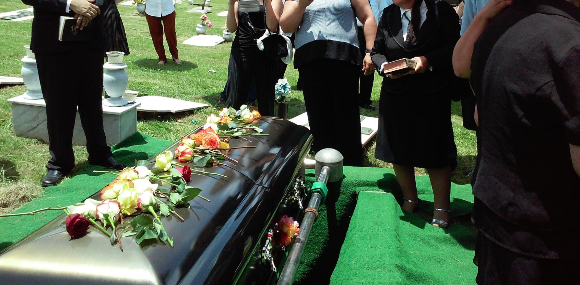 a black casket sitting on top of a grave is covered with roses. The peopel standing around the casket are wearing black clothing. Some people are holding books which could be bibles. The gravesite is covered with astroturf.
