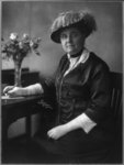 Jane Addams sits at a desk in front of a bouquet and holds a pen. She wears a hat and gloves.