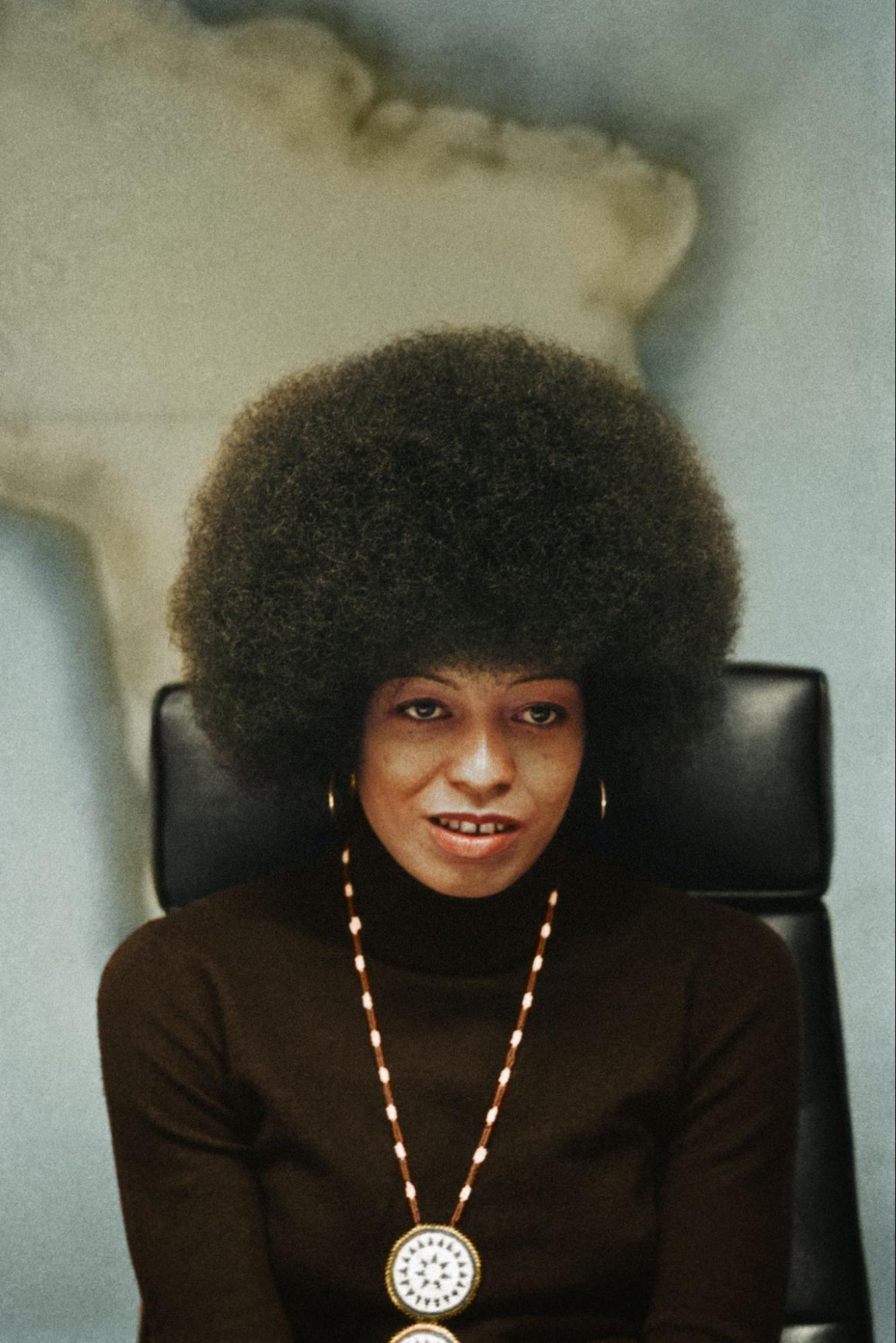 In a photo from the 1970s, Angela Davis sits in front of a map of Africa and wears a necklace that contrasts with her black tutleneck.