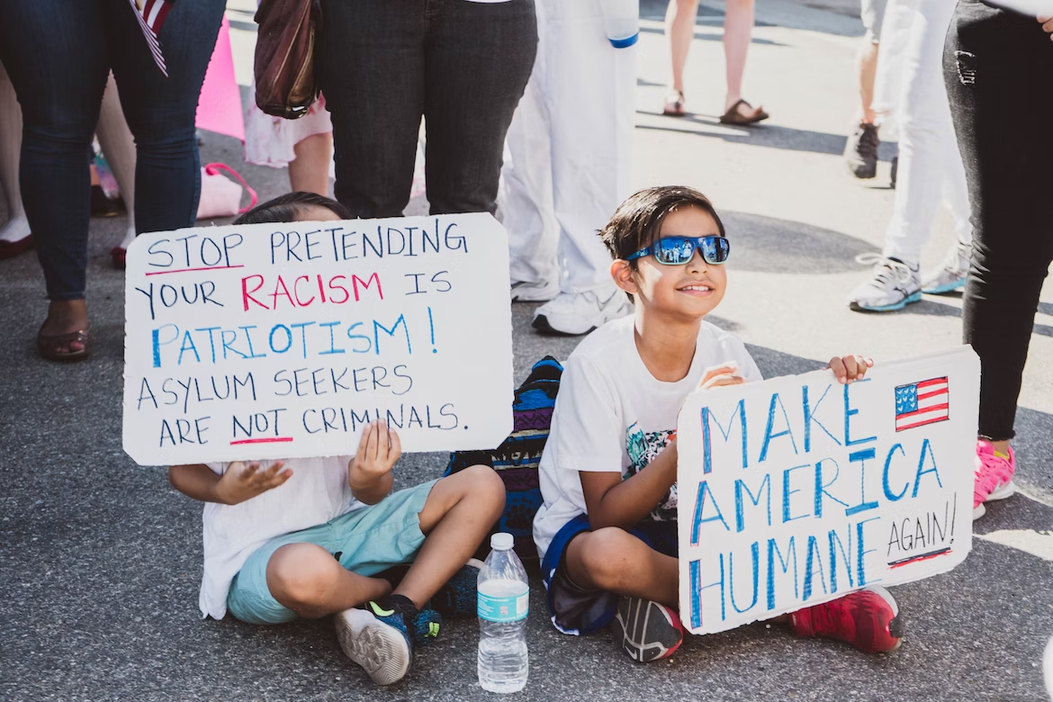 Two children smile and hold signs that say, "Make America humane again" and "Stop pretending your racism is patriotism! Asylum seekers are not criminals."