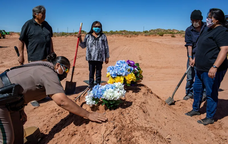 Five Indigenous people hold shovels in a circle around a fresh mound of direct decorated with colorful flowers