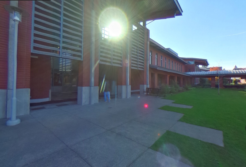 The modern two story brick building has a concrete patio and a grassy area surrounding it. You can also see a covered walkway and a roof that protects the patio.