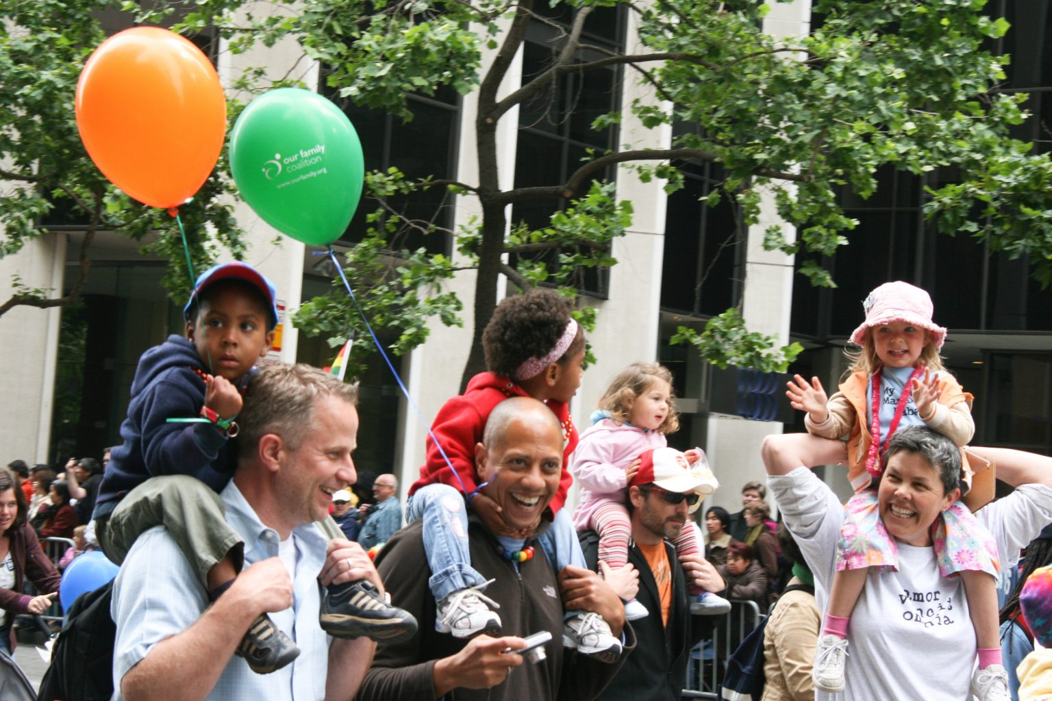Parents with children on their shoulders smile, hold balloons, and walk down the street