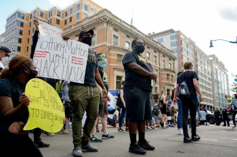 A street has demonstrators standing, kneeling, and walking by. One person holds a sign that reads, "Nothing matters until Black lives do."