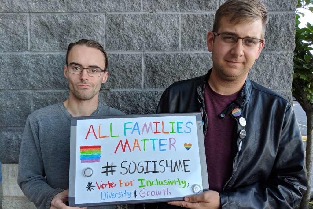 Two men hold a colorful sign reading, "all families matter #sogiis4me, vote for inclusivity, diversity, and growth"