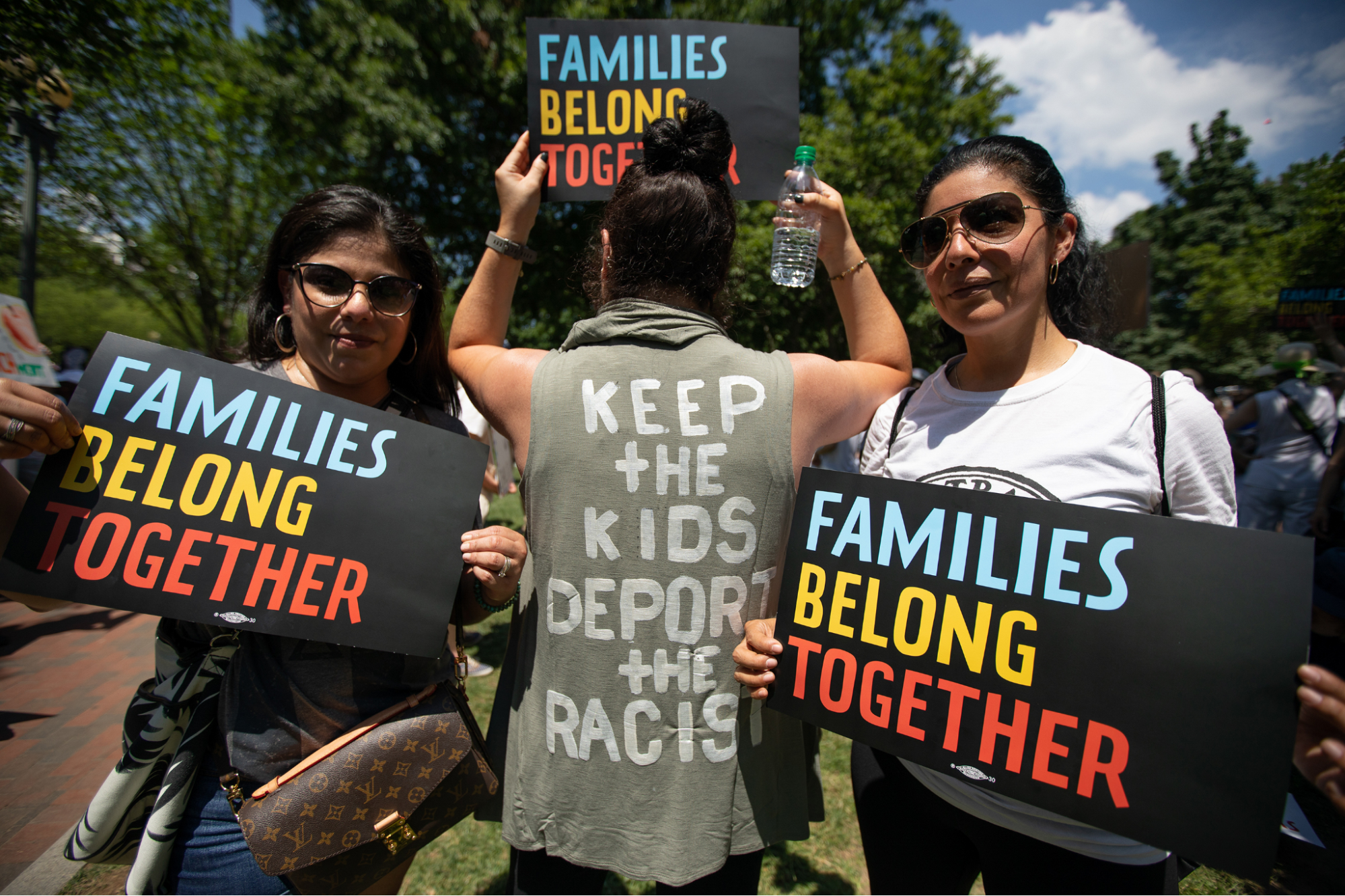 Three people with signs a t-shirts reading, "families belong together" and "keep the kids, deport the racists"