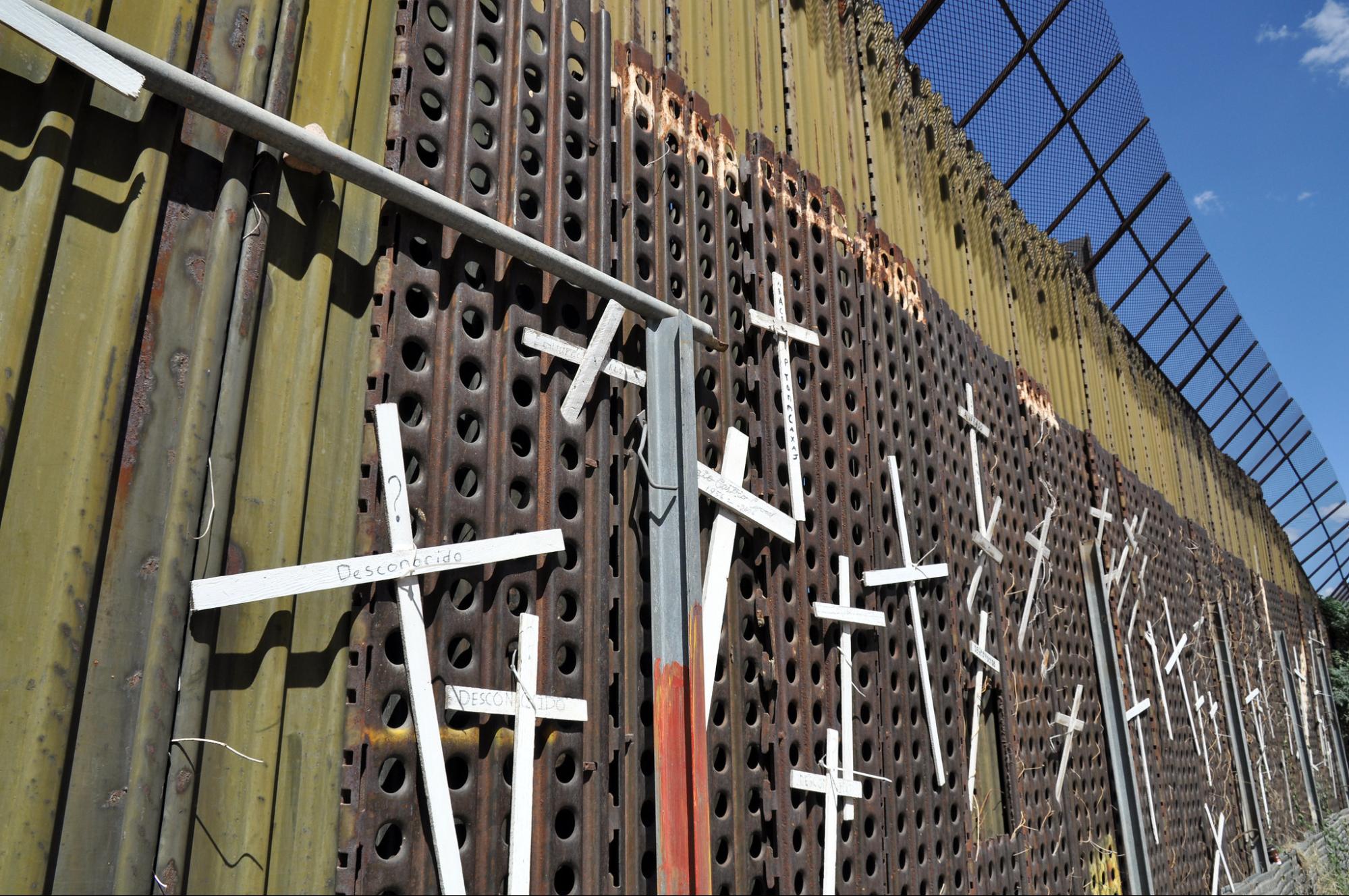 A wall made of metal sheeting and topped with wire extends into the distance. Simple wooden crosses cover the wall. Although the writing is almost invisible, the names of people who have died crossing the border are written on them.