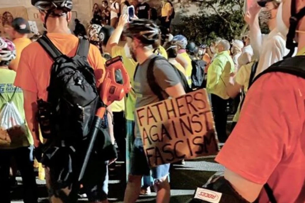We see the back of one man with a backpack and leaf blower, wearing a helmet. Another man wearing a build holds a sign that says"fathers against fascism." They are surrounded by many other men who are protesting.