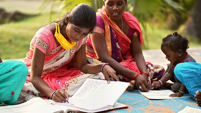 Two women in pink saris sit on a blanket with a toddler, writing in large notebooks.