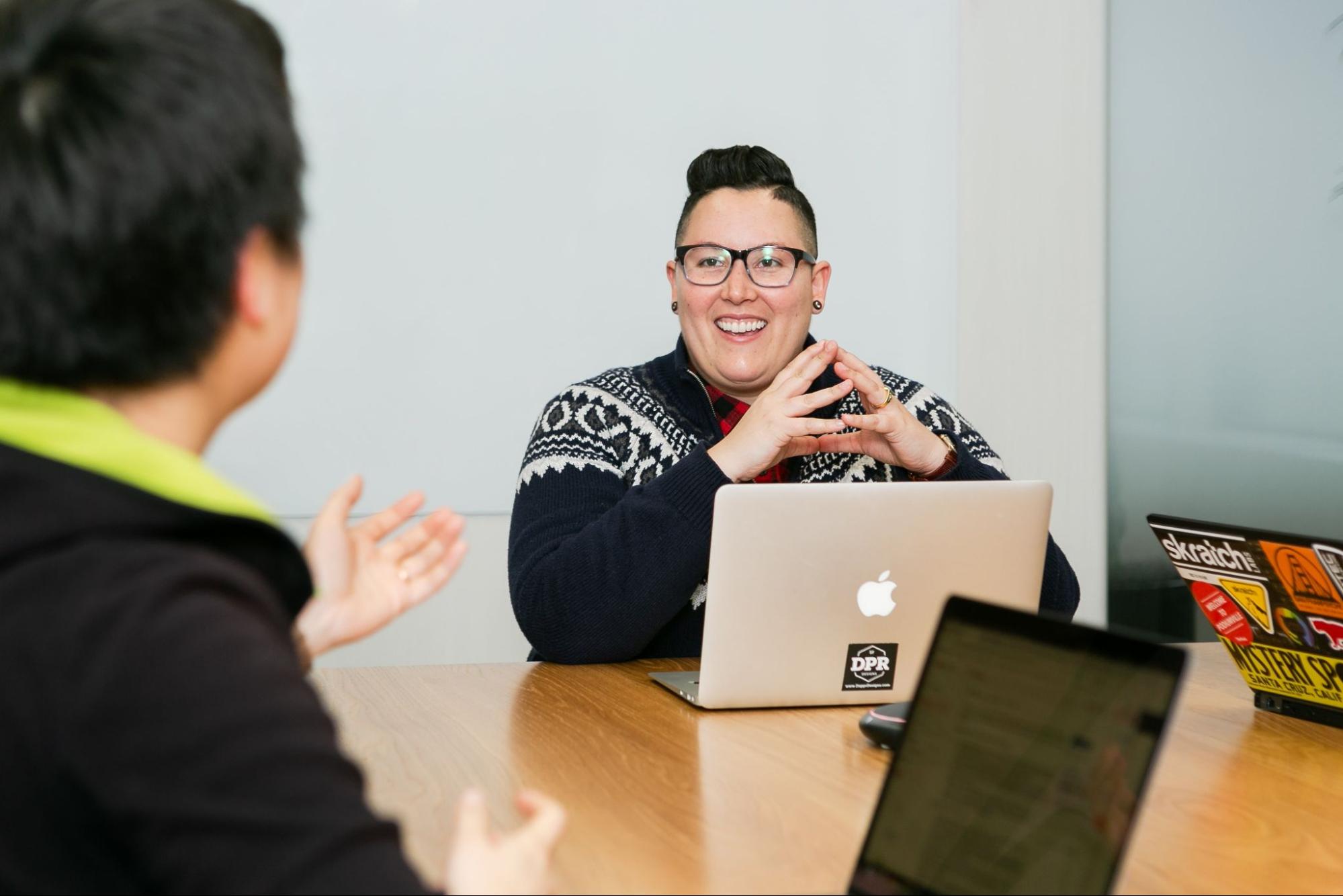 Gender fluid person sits in front of a laptop smiling at a person across the table