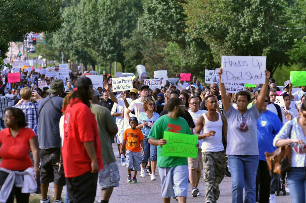 A crowd of people of diverse ages and ethnicities march down a treed street holding signs like "Hands up don't shoot"