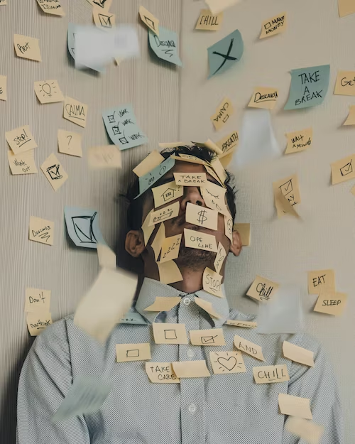 A person wearing a blue shirt is standing in a corner. He is covered by post its. The wall is covered by post its too. The post its say things like "take a break", "Chill" "heart" "Money" "Checkbox" and "Stop".