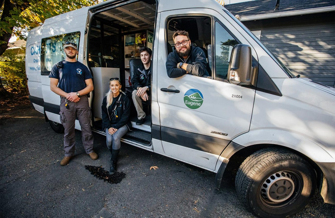 Four people in blue shirts standnear and sit in a white van, with a logo for Eugene Oregon.