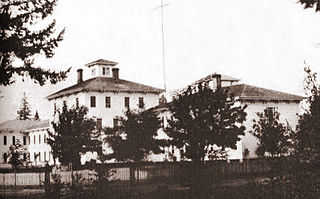 the old photograph shows two or three large white buildings of two or three stories. They look like they are made of wood. In the foreground there are trees. A fence surrounds the buildings, land and trees.