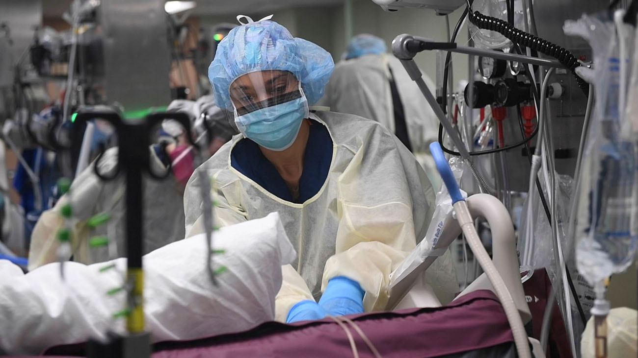 Medical provider wears protective gear. Their mouth and nose is covered by a mask, and their face is covered by a shield. They are helping a patient on a bed in front of them.