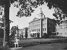 The image shows a huge brick building. The building has four stories and a cupola. In the foreground there is a large tree, with a woman swinging on a swing, attached to the large tree.