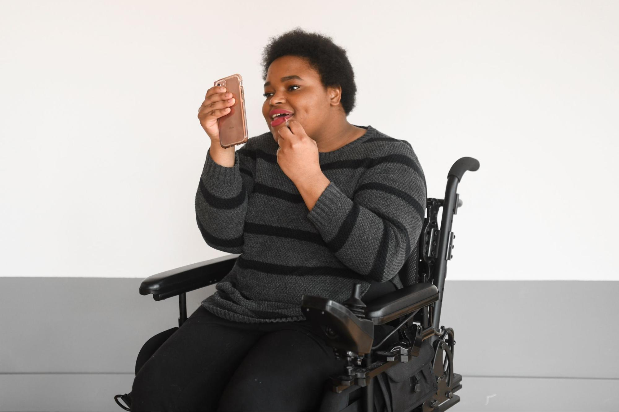 Torso crop of a Black non-binary person applying pink lip gloss by using their pink cell phone as a mirror. They are sitting in a power wheelchair and in front of a white wall with a gray stripe.