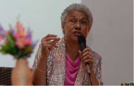 Patricia Hill Collins gestures with one hand and holds a microphone with another. She looks earnest and wears glasses and a patterned cardigan.