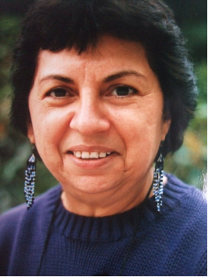 Gloria Anzaldúa wears beaded earrings and a blue sweater. She smiles directly at the photographer.