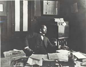 W.E.B DuBois looks up from a desk covered with newspapers and books to look out the window thoughtfully