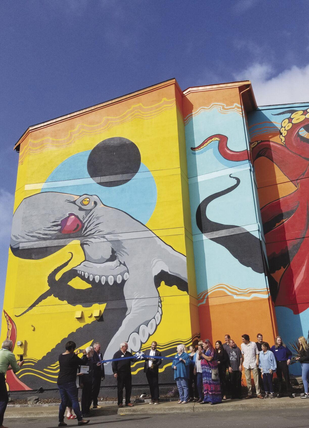 Men in suits hold a ribbon cutting ceremony in front of a mural on a building. People watch and take pictures.