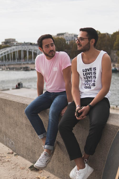 Two men wearing jeans, sneakers, and t-shirts sit on a wall. One t-shirt reads "love is love is love is love is love is love" in English.