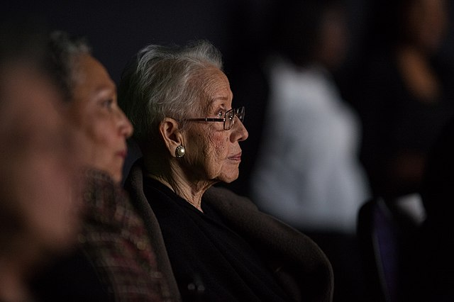 Katherine Johnson, seen from the side, sits in a dark movie theater amongst over moviegoers, her face lit by the screen