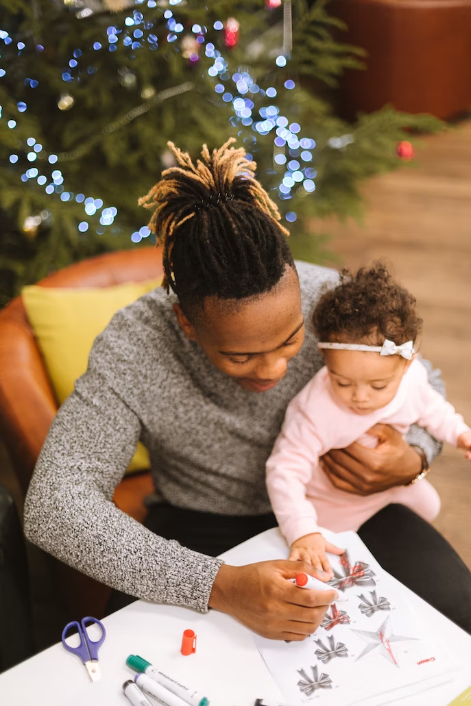 A Black father holds his infant child on his lap while they color in front of a decorated holiday tree