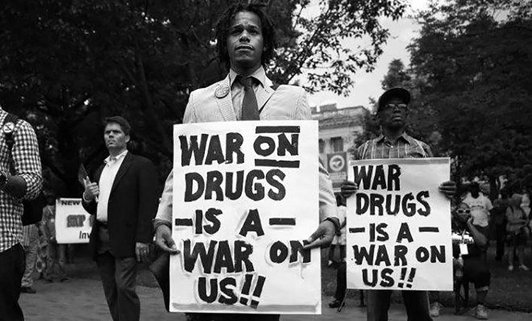 a young man with dark skin wearing a suit and tie is holding a protest sign that says, " War on Drugs is a War on us!!"