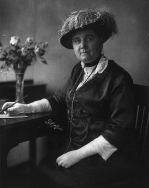 Jane Addams sits at a desk in front of a bouquet and holds a pen. She wears a hat and gloves.