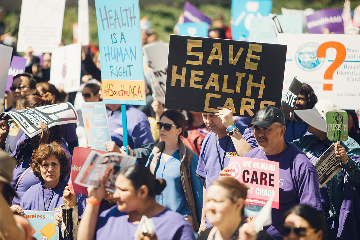 Fig 7.18 A large group of people gather at a rally. They hold signs reading Health is a Human Right and Save Health Care.