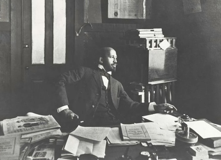 W.E.B. DuBois looks up from a desk covered with newspapers and books to look out the window thoughtfully.