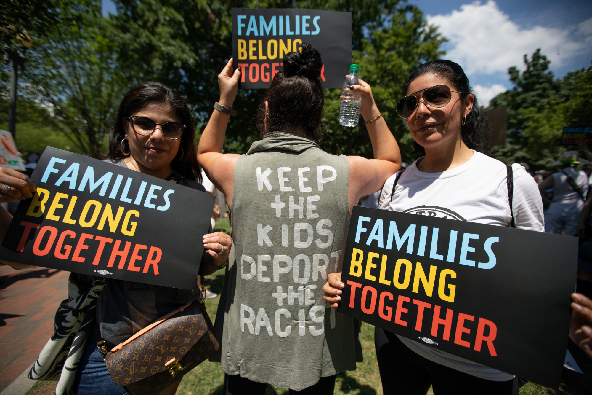 Three people with signs a t-shirts reading, &quot;families belong together&quot; and &quot;keep the kids, deport the racists&quot;