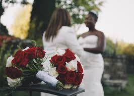 Two bouquets of flowers in sharp focus; behind the flowers in soft focus: two women in white wedding themed clothing, a White woman in a suit and a Black woman in a dress.