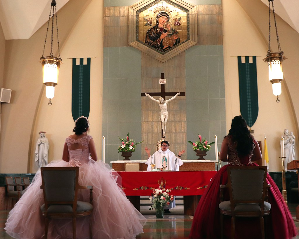 Two girls in quinceanera dresses facing the altar and priest at a Catholic church.