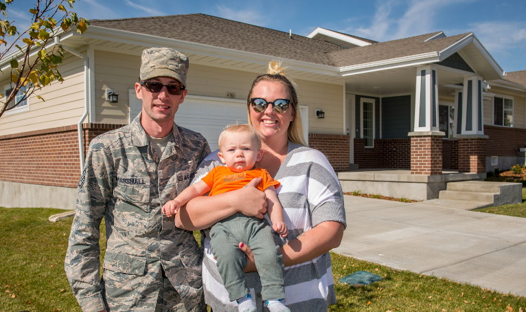 A military couple and their toddler smiling in front of their home.