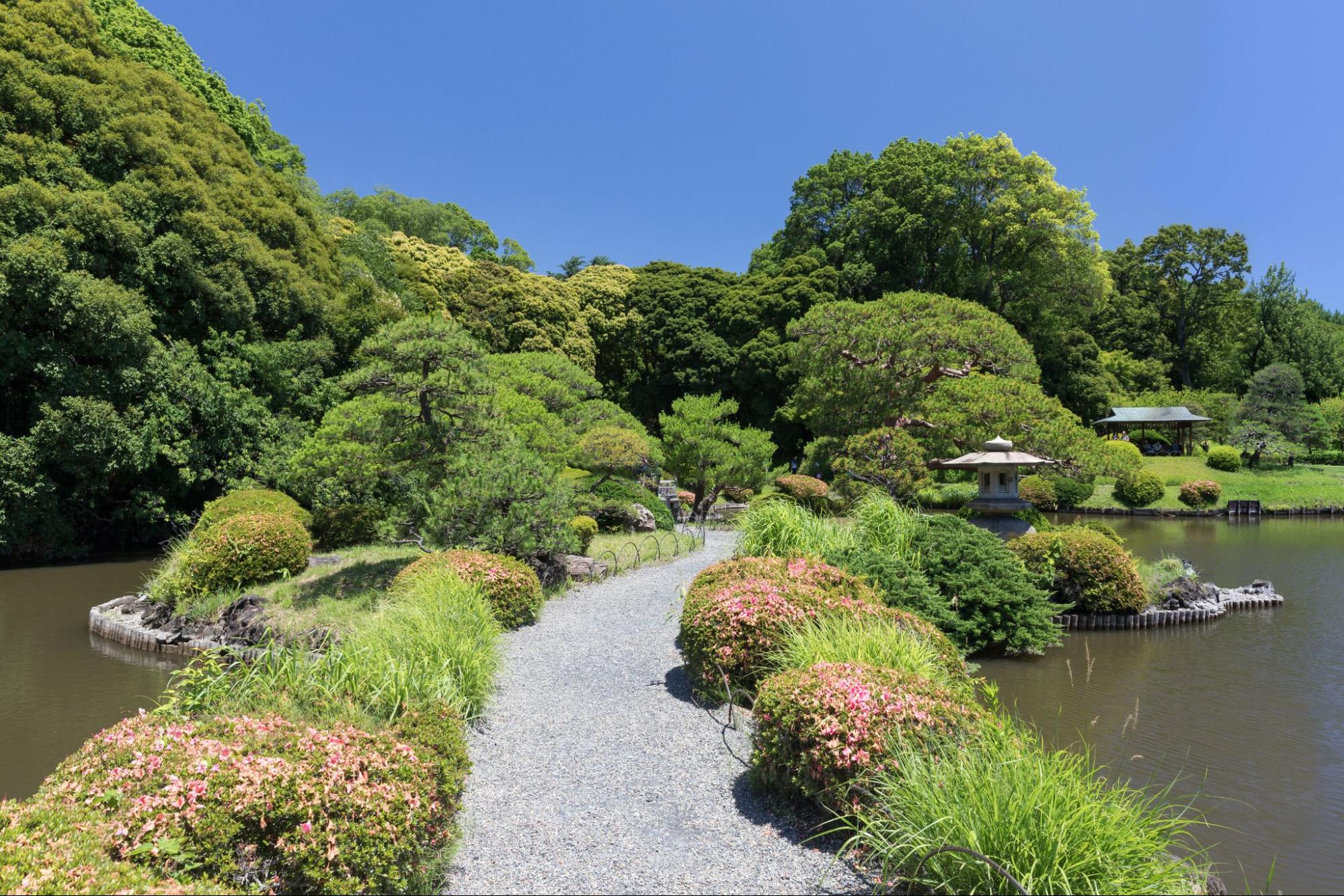 A gravel path between two small lakes, leading to a Japanese-style garden with trees, bushes and small stone pagodas