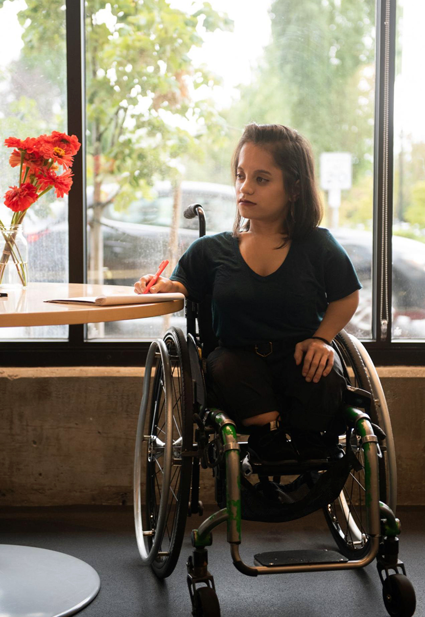 A South Asian person in a wheelchair looks thoughtfully to the side while taking notes. She is backlit by the window she's in front of and wearing a teal shirt with black pants and shoes.