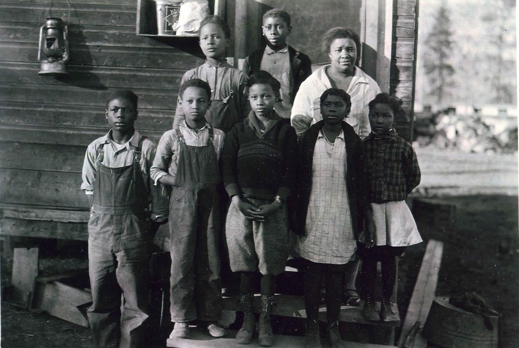 The image shows a black and white photograph of eight individuals. In the foreground, seven children of varying ages stand on a small wooden step in front of a wooden structure, possibly a house. They are dressed in a mix of overalls, dresses, and sweaters, typical of early to mid-20th century rural attire. The children are positioned in two rows, with the tallest in the back. Behind them, an adult woman stands, wearing a light-colored blouse. Behind them appears to be the exterior of a small, log school house.
