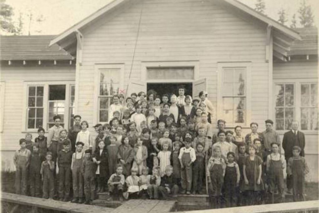 The image is a vintage black-and-white photograph depicting a large group of children and adults posed in front of a large wooden schoolhouse. The building is a classic, early 20th-century structure with clapboard siding, several windows with multiple panes, and a gabled roof. The group consists of approximately 30 children of various ages standing in several rows. The children are dressed in period attire, with many boys wearing overalls and girls in dresses. A few teachers stand amidst the group.