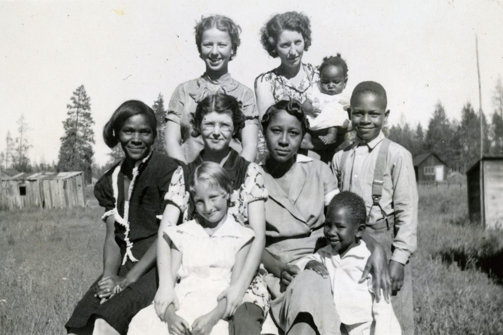 A group of both Black and White children of various ages, from toddler to teenager, are posed together for a school photograph. One white girl standing in the back is holding a Black toddler in her arms.