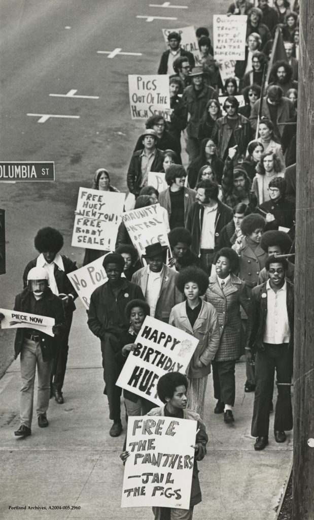 Protestors marched from Portland State University to the Federal Courthouse in support of community-controlled policing. The image shows the protestors, with signs, marching down the sidewalk outside the courthouse.