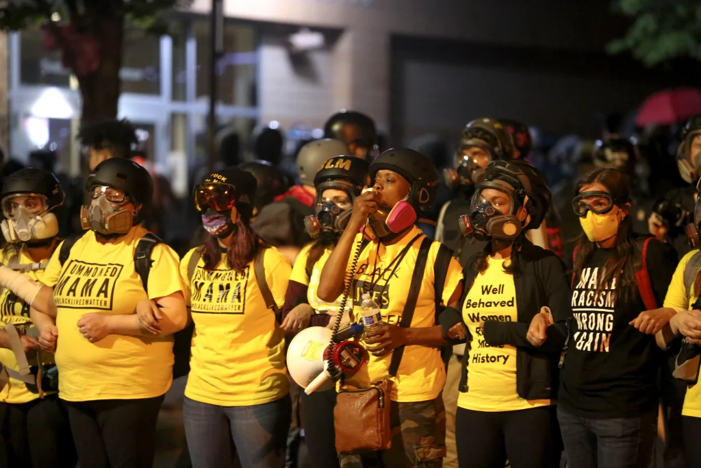 A wall of mothers from Mxm Bloc in yellow shirts standing at the front of a protest against racial injustice and police brutality. They have their arms linked together and many are wearing either eye protection or gas masks.