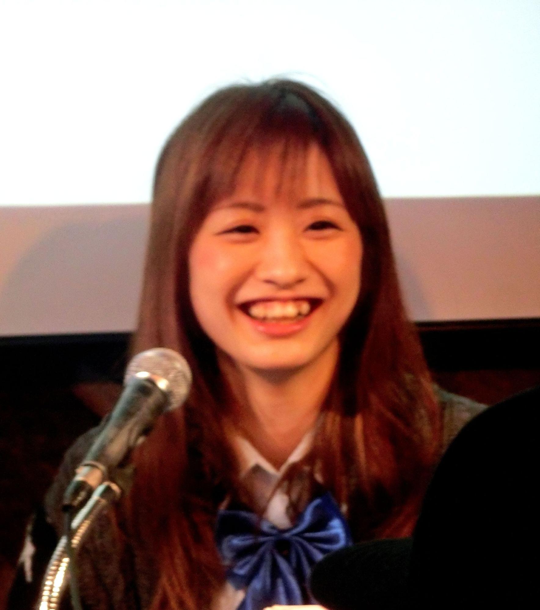 An Asian woman with a pronounced canine tooth smiles in front of a microphone.