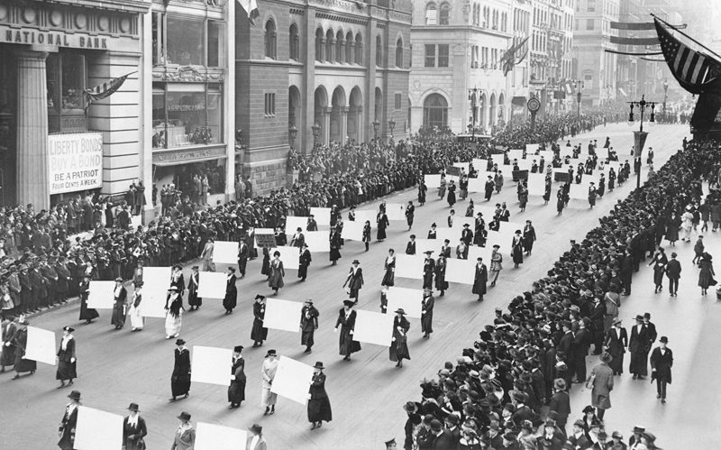 Women marching in the middle of a street in New York city in 1917 at a Women's suffragist parade. Crowds of people line the streets and American flags are flying at local businesses. A bank is advertising "be a patriot by getting bonds."