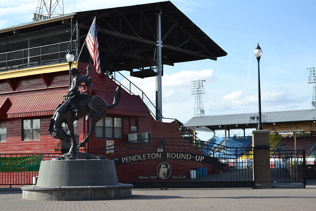 Pendleton Round-Up Since 1910: Let 'er Buck! Statue in front of gates of a man riding a horse. American flag visible on building behind statue.